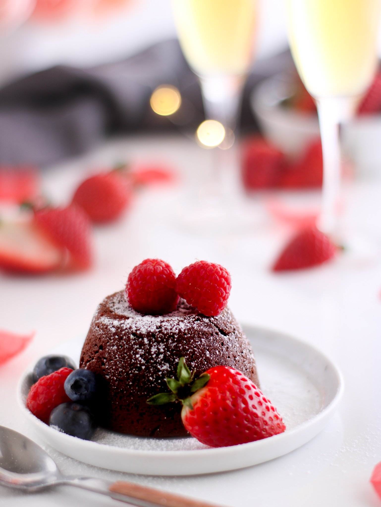 Gluten-free chocolate molten lava cake sitting on a white plate surrounded by fresh strawberries, raspberries and blueberries with two glasses of champagne in the background.